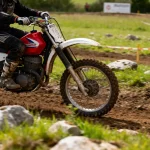 Motocross en plein air, moto rouge et blanche, pilote en tenue noire, terrain boueux avec des rochers.