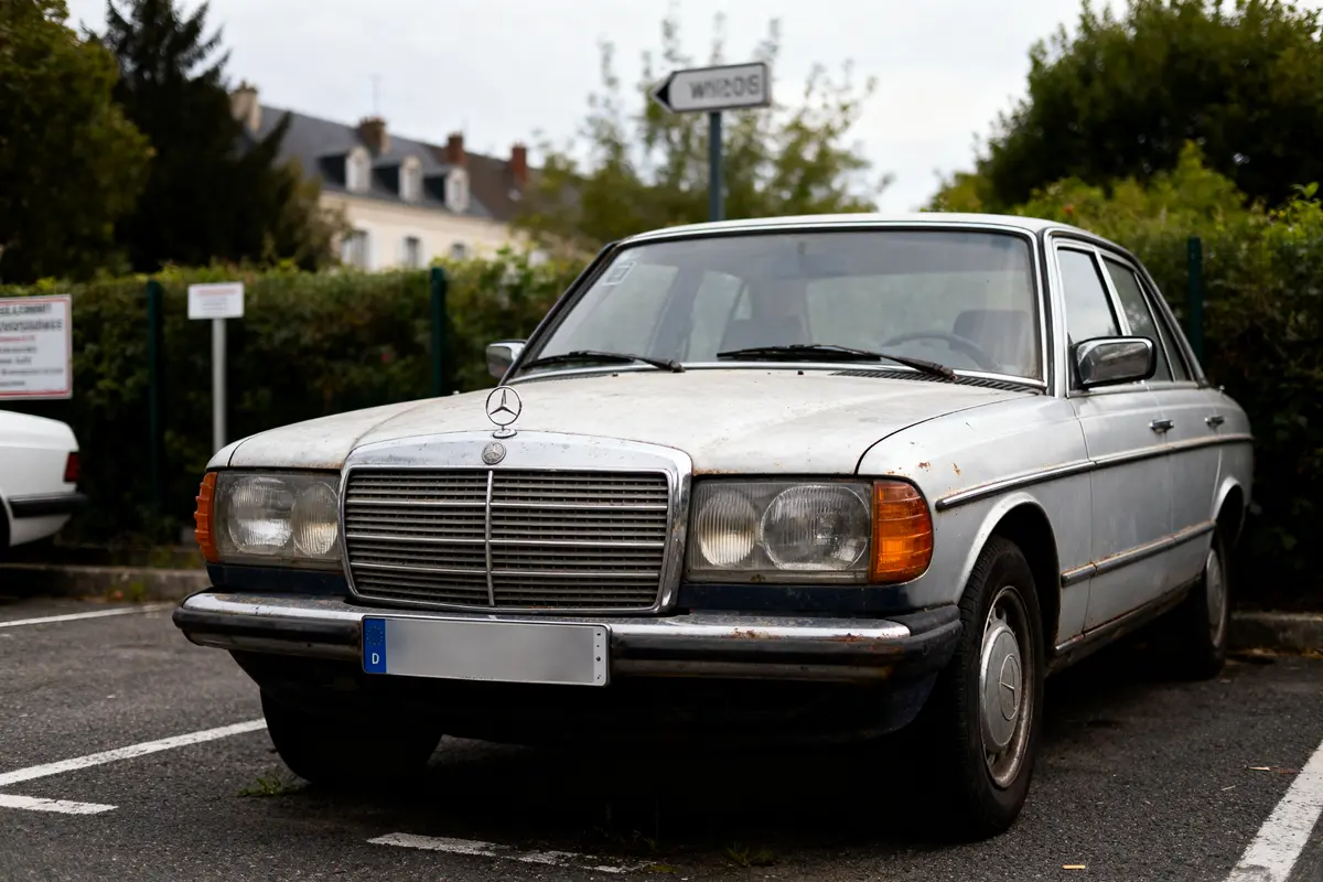Voiture ancienne Mercedes grise stationnée dans un parking, bâtiments et arbres en arrière-plan.