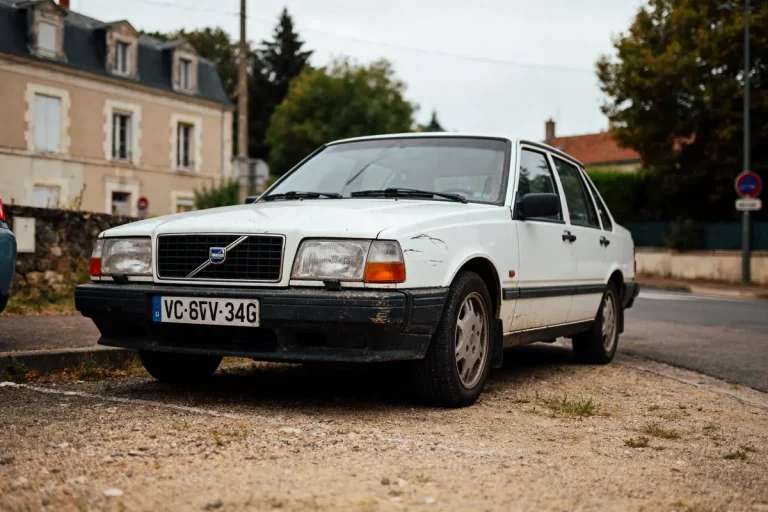 Voiture blanche vieille, stationnée sur un trottoir, devant une maison ancienne, avec végétation en arrière-plan.
