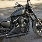 Moto noire customisée garée sur la route, moteur visible, pneus épais, réservoir marqué d'un logo.