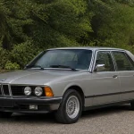 Voiture classique grise stationnée, modèle BMW 733i, avec des phares ronds et environnement arboré